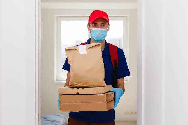 Empleado de repartidor en guantes de máscara uniforme de camiseta de gorra roja dan cajas de pizza de pedido de comida aisladas en el estudio de fondo amarillo. Servicio de cuarentena pandemia coronavirus virus gripe 2019-ncov concept