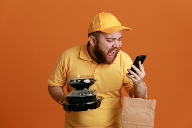 Foto empleado repartidor con uniforme de camiseta en blanco con gorra amarilla sosteniendo contenedores de comida con una bolsa de papel hablando por teléfono móvil enojado y frustrado gritando y gritando de pie sobre fondo naranja