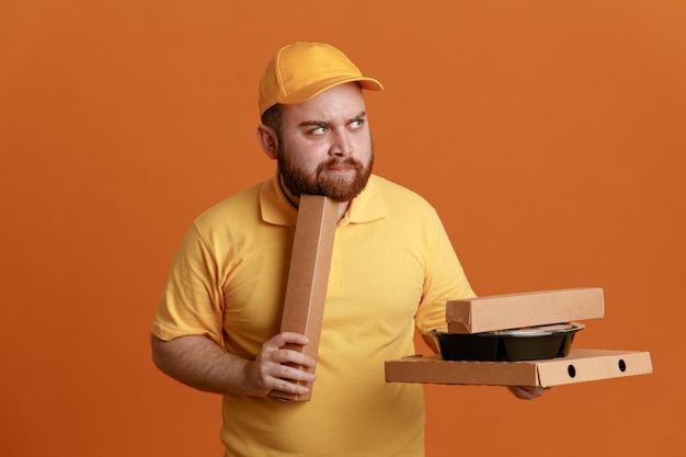 Empleado repartidor en uniforme de camiseta en blanco con gorra amarilla sosteniendo contenedores de comida y caja de pizza mirando a un lado con expresión pensativa pensando de pie sobre fondo naranja