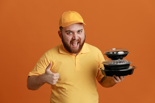 Empleado del repartidor con uniforme de camiseta en blanco con gorra amarilla sosteniendo contenedores de comida mirando a la cámara feliz y emocionado sonriendo ampliamente mostrando el pulgar hacia arriba de pie sobre fondo naranja