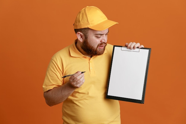 Empleado repartidor en uniforme de camiseta en blanco con gorra amarilla sosteniendo portapapeles y bolígrafo mirando el portapapeles confundido de pie sobre fondo naranja