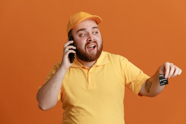 Empleado repartidor en uniforme de camiseta en blanco con gorra amarilla con tarjeta de crédito hablando por teléfono móvil de pie feliz y positivo sobre fondo naranja