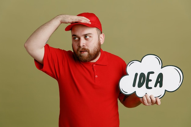 Empleado repartidor con uniforme de camiseta en blanco con gorra roja con discurso de burbuja con idea de palabra mirando a lo lejos con la mano sobre la frente de pie sobre fondo verde