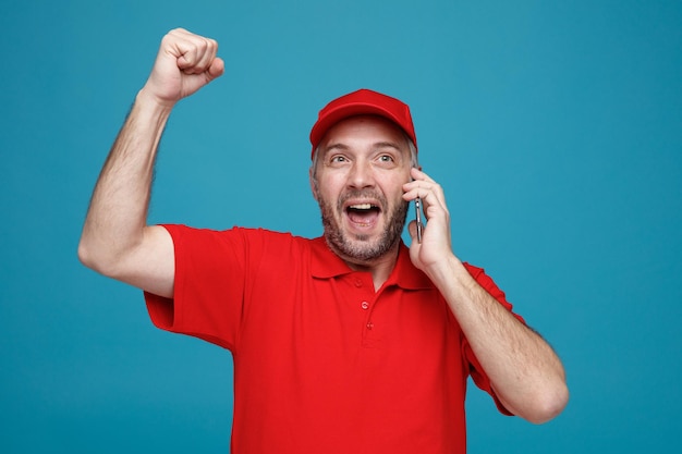 Foto empleado repartidor en uniforme de camiseta en blanco con gorra roja hablando por teléfono móvil loco feliz y emocionado levantando el puño cerrado de pie sobre fondo azul