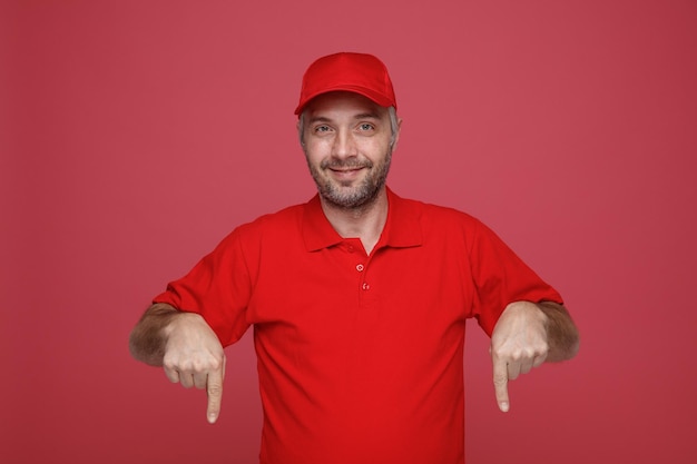 Empleado repartidor en uniforme de camiseta en blanco con gorra roja mirando a la cámara sonriendo feliz y positivamente señalando con los dedos índices hacia abajo de pie sobre fondo rojo