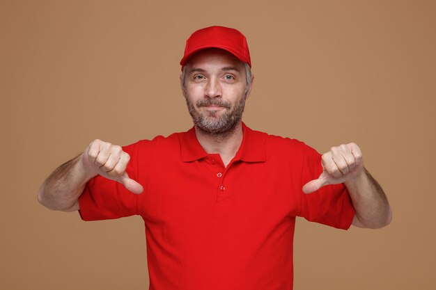 Empleado repartidor en uniforme de camiseta en blanco con gorra roja mirando a la cámara sonriendo satisfecho de sí mismo de pie sobre fondo marrón