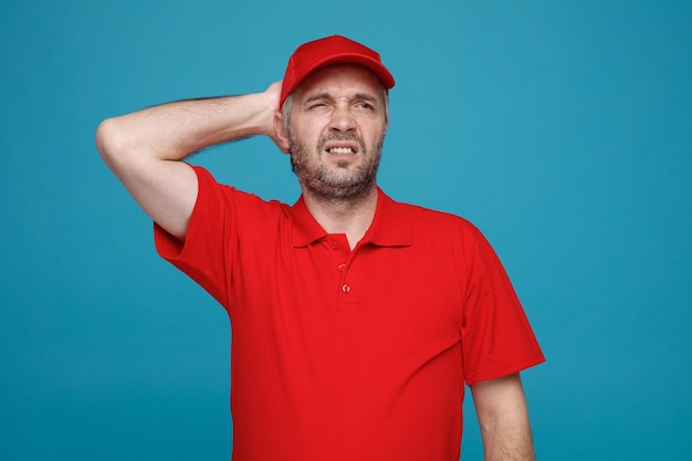 Empleado repartidor en uniforme de camiseta en blanco con gorra roja mirando a un lado confundido sosteniendo la mano en su cabeza por error de pie sobre fondo azul