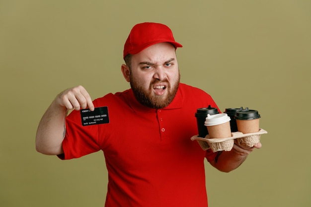 Empleado repartidor en uniforme de camiseta en blanco con gorra roja sosteniendo tazas de café mostrando su tarjeta de crédito esperando el pago con cara enojada de pie sobre fondo verde