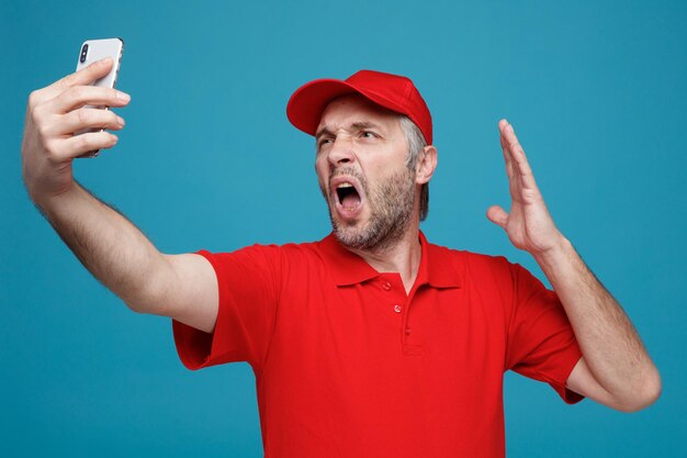 Empleado repartidor en uniforme de camiseta en blanco con gorra roja sosteniendo un teléfono inteligente con videollamada enojado y frustrado gritando de pie sobre fondo azul