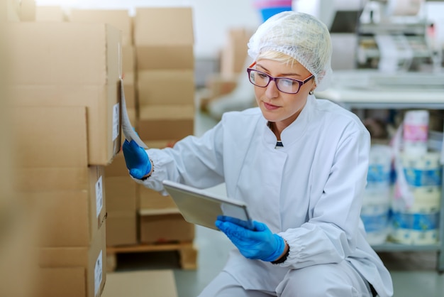 Empleado rubio caucásico en uniforme estéril agachado junto a cajas y lidiando con logística de productos. Interior de la fábrica de alimentos. Tableta en mano.