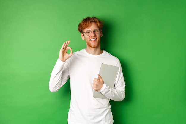 Empleado de sexo masculino joven hermoso en vidrios que muestran el signo de Ok, sosteniendo la computadora portátil, colocándose sobre fondo verde.