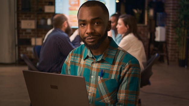 Empleado sonriente afroamericano trabajando en retrato de laptop, cámara lenta, vista frontal. Trabajador de oficina sosteniendo una computadora portátil, mirando la cámara de tiro medio, reunión de negocios en segundo plano.