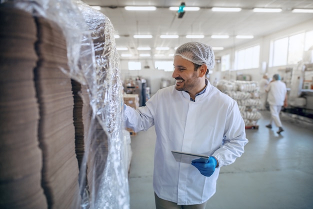 Foto empleado sonriente caucásico en uniforme blanco estéril con tableta en fábrica de alimentos.