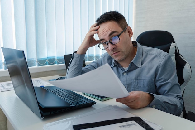 Un empleado se sorprende al leer un documento en papel mientras está sentado en el lugar de trabajo Empresario deprimido por la contabilidad en su oficina