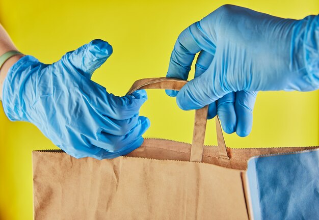 El empleador del hombre de entrega en guantes azules sostiene la bolsa de papel del arte con la comida, aislada en estudio amarillo. Servicio de cuarentena del virus del coronavirus pandémico concepto 2019-ncov.