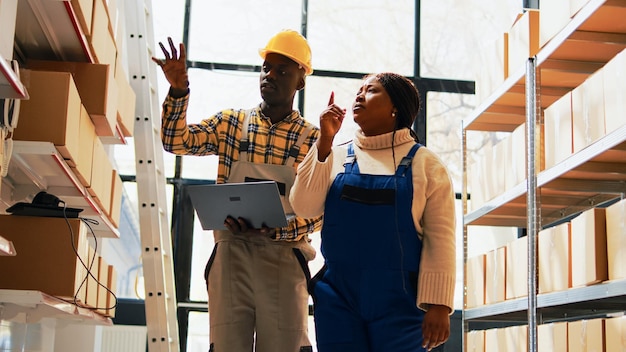 Empleados afroamericanos que usan una computadora portátil en la sala de almacenamiento, hombres y mujeres que revisan la mercancía y el almacén en el depósito. Equipo de trabajadores analizando logística en pc. Disparo de mano.