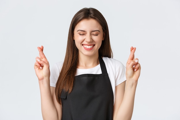 Empleados, empleo laboral, pequeñas empresas y concepto de cafetería. Esperanzado barista lindo emocionado y optimista en delantal negro pidiendo deseos, cruce los dedos, buena suerte mientras reza.