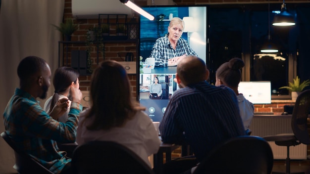 Foto empleados saludando a un compañero de trabajo adulto medio en una videollamada, saludando a sus colegas, comunicación por videoconferencia. trabajadores de oficina, escuchando hablar a una mujer de negocios, hablando en teleconferencia por la noche