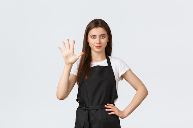 Empleados de la tienda de abarrotes, concepto de pequeñas empresas y cafeterías. Alegre joven barista en delantal negro mostrando el número cinco, sonriendo determinado, trabajando en el café