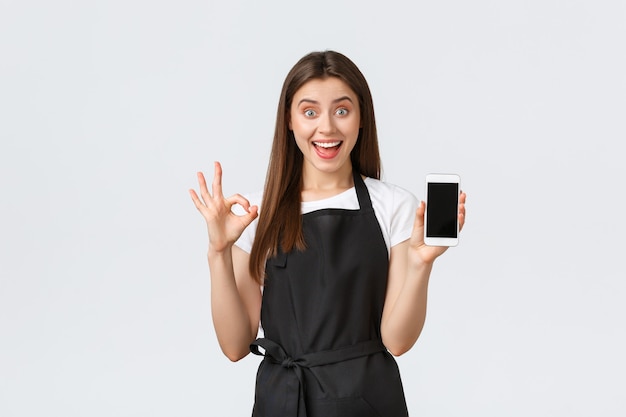 Empleados de la tienda de abarrotes, concepto de pequeñas empresas y cafeterías. La barista femenina complacida y emocionada con delantal negro garantiza una buena experiencia de usuario que muestra la aplicación de pantalla del teléfono inteligente, haga el signo correcto.