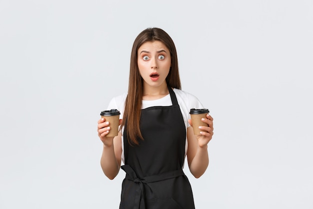 Empleados de la tienda de abarrotes, concepto de pequeñas empresas y cafeterías. Barista joven preocupada hizo un pedido incorrecto, sosteniendo dos tazas de bebidas y jadeando sorprendido, fondo blanco.