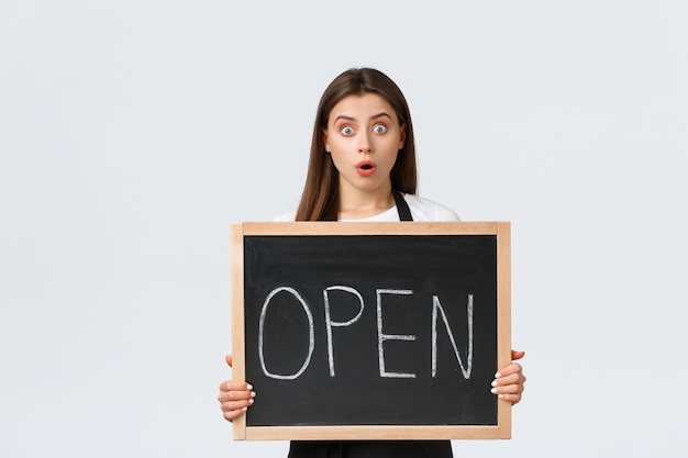 Empleados de la tienda de abarrotes, concepto de pequeñas empresas y cafeterías. La cámara de la mirada de la muchacha sorprendida con la boca abierta informa a los clientes que la tienda está abierta, que muestra la muestra, fondo blanco de pie.