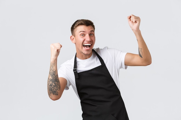 Empleados, tiendas de abarrotes y concepto de cafetería. Alegre trabajador de la tienda de abarrotes, barista con delantal negro regocijándose con las maravillosas noticias, diciendo que sí, ganando, levantando las manos en señal de triunfo