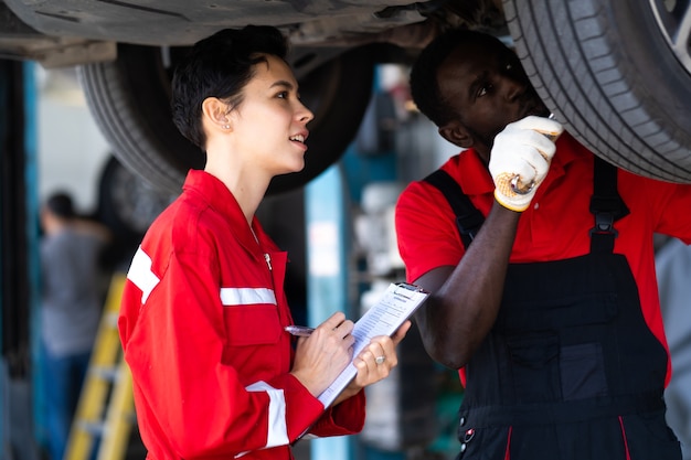 Empoderamiento de waman caucásico mecánico con uniforme rojo trabajando debajo de un vehículo en una estación de servicio de automóviles