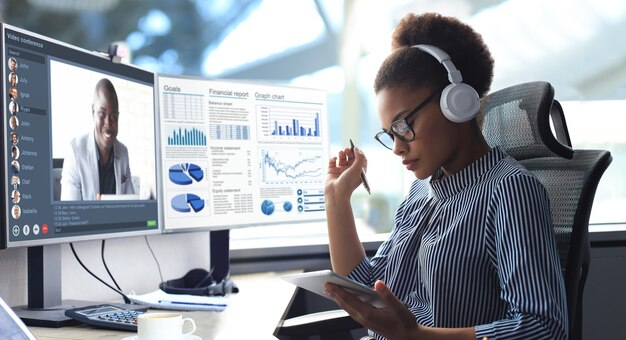 Empreendedor afro-americano usando fones de ouvido, conversando com seus colegas em videoconferência. Equipe multiétnica de negócios trabalhando no escritório usando o PC, discutindo o relatório financeiro da empresa.