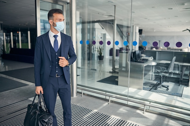 Empreendedor atraente segurando uma mala de couro e em pé com uma máscara médica no rosto ao sair do aeroporto. Banner modelo