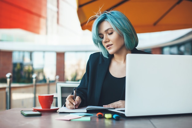 Empreendedor caucasiano com cabelo azul, trabalhando em uma lanchonete no laptop e fazendo algumas anotações em um livro enquanto bebe um café