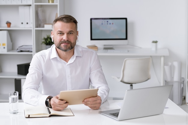 Empreendedor confiante com tablet e airpods sentado na mesa e olhando informações online no escritório