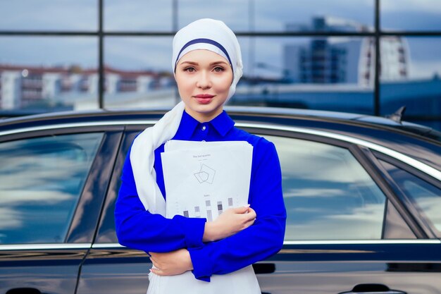 Empreendedor de mulher de negócios muçulmano europeu bonito e bem sucedido em um elegante hijab e turbante segurando o papel de arranha-céu fundo de janelas de escritório em pé de rua perto de seu carro preto