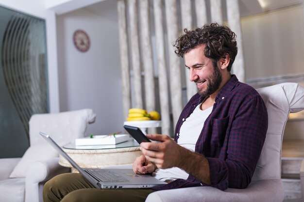 Empreendedor de sucesso sorrindo de satisfação enquanto verifica as informações em seu laptop enquanto trabalha em um escritório doméstico. jovem relaxando no sofá com um laptop e um celular nas mãos