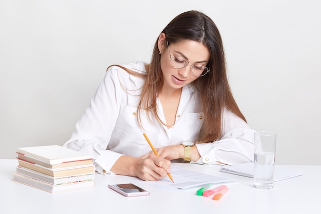 Empreendedor feminino escreve plano de organização, senta-se na área de trabalho, usa livros e lápis, bebe água fresca de vidro, concentrado no trabalho, usa óculos ópticos redondos, isolados no branco