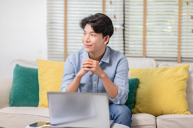 Empreendedor homem de negócios asiático bonito vestindo camisa sentado no sofá trabalhando on-line com computador portátil sorrindo felicidade e relaxar no escritório em casa Homem jovem usando mídias sociais Pequeno negócio Startup