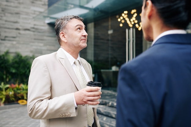 Empreendedor idoso pensativo tomando café com seu colega de trabalho e olhando para o prédio em que está trabalhando