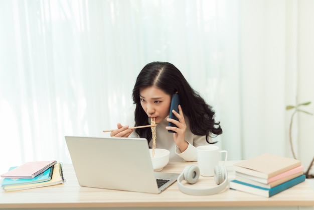 Empreendedor jovem asiático sorridente confiante na hora do almoço enquanto está sentado na mesa do escritório e usando o telefone celular