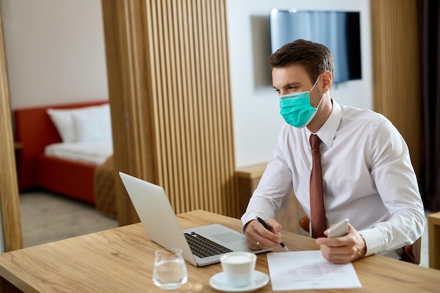 Empreendedor masculino com máscara facial usando laptop enquanto trabalhava em relatórios de negócios no quarto de hotel