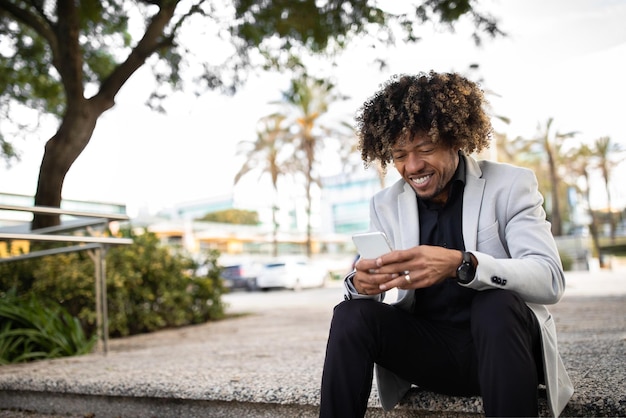 Empreendedor negro bem-sucedido de meia-idade usando mensagens de texto no celular ou navegando na Internet sentado