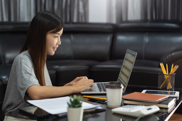 Empreendedor ou freelancer asiático bonito novo atrativo da mulher que trabalha em casa com relatórios comerciais do portátil e comunicações online no sofá da sala de visitas, trabalhando remotamente acessa o conceito.