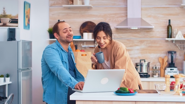 Empreendedor trabalhando no laptop na cozinha e a esposa segurando uma xícara de café, olhando para o trabalho dele. marido e mulher cozinhando comida de receita. estilo de vida feliz juntos e saudável. família em busca de refeição online