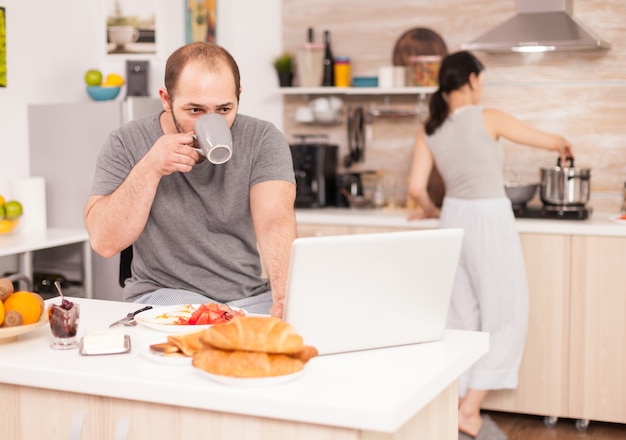 Empreendedor trabalhando no laptop na cozinha e bebendo café enquanto a esposa está preparando o café da manhã. Freelancer trabalha online através da Internet usando tecnologia digital moderna