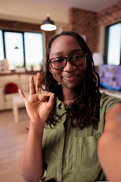 Foto empreendedor trabalhando remotamente fazendo sinal de ok na frente da câmera em videoconferência na sala de estar de casa. mulher afro-americana sorrindo para a câmera fazendo gesto com a mão enquanto conversava com os colegas.