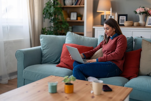 Empreendedora focada em roupas casuais de negócios fazendo pesquisa em laptop enquanto relaxa no sofá na sala de estar