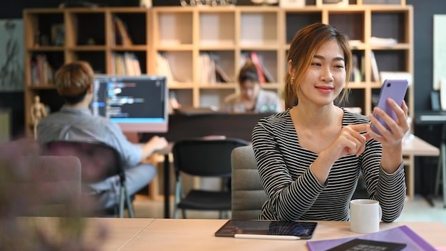 Empregada asiática sorridente sentada no escritório da caixa e usando telefone celular