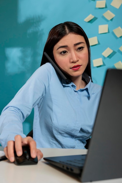 Empregada corporativa falando com colega ao telefone enquanto fazia uma pesquisa de informações no software corporativo. Mulher profissional usando computador portátil na mesa de escritório.