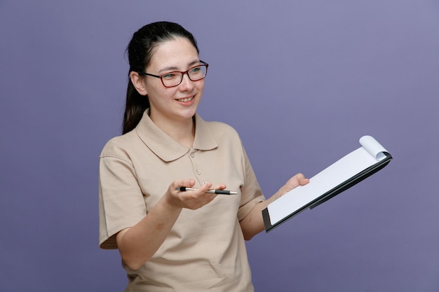 Empregada de entrega em uniforme de camisa polo em branco usando óculos segurando prancheta e caneta parecendo confiante sorrindo feliz e positivo em pé sobre fundo azul