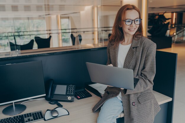 Empregada de escritório sorridente, sentada na mesa em seu local de trabalho, segurando um laptop