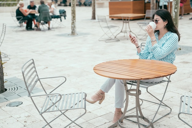 Empregada de mulher jovem e bonita em óculos de sol e roupa casual inteligente, sentado na loja de café ao ar livre de mesa na rua. senhora de escritório elegante aproveite a hora do almoço usando telefone celular e bebendo café.